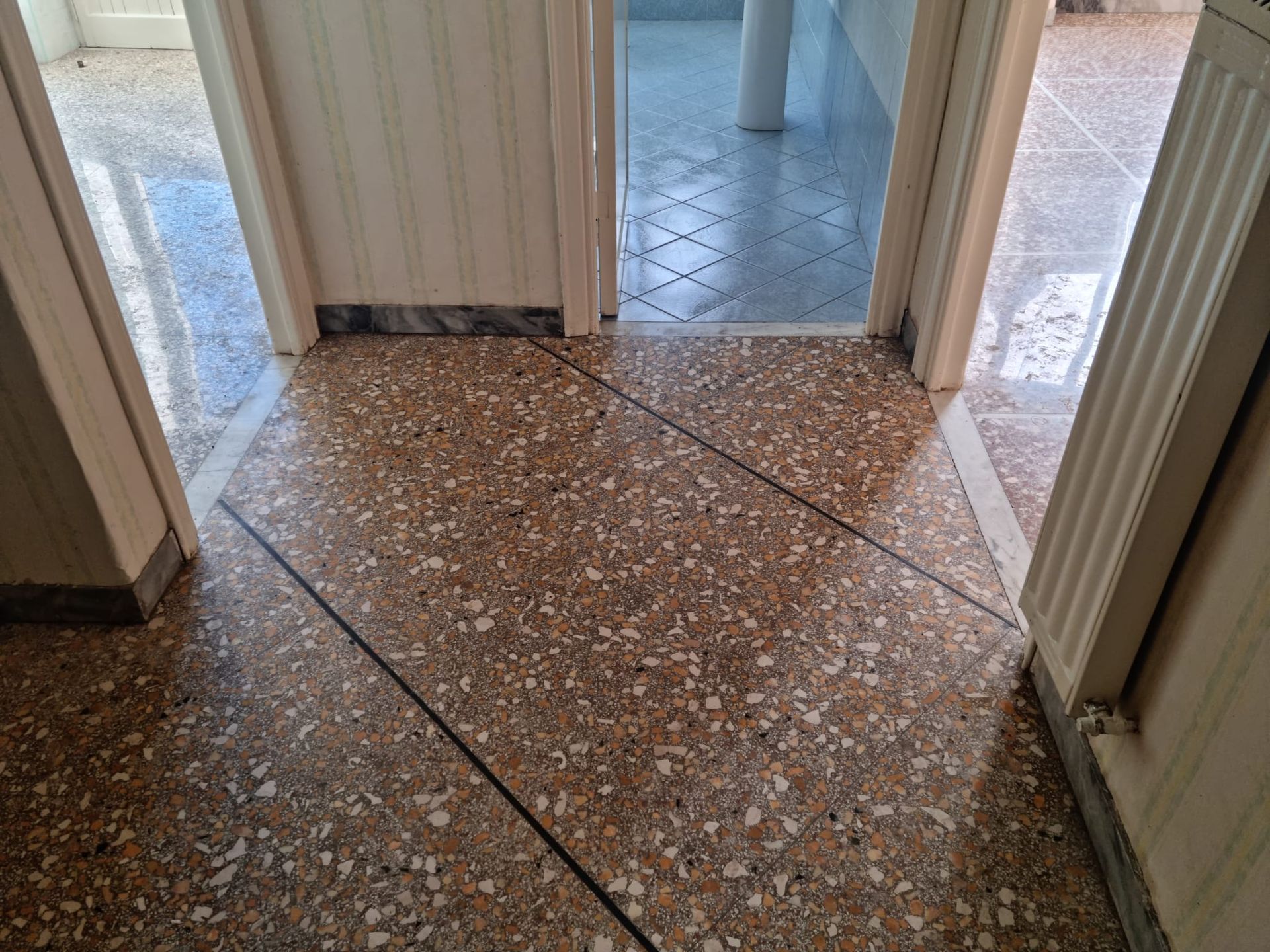 Corridoio piastrellato con pavimenti a macchie beige. Due porte conducono ad altre stanze con pavimenti diversi.