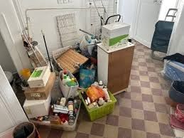 Stanza disordinata con varie scatole, cestini e oggetti ammucchiati sul pavimento; sono presenti anche un pavimento a scacchi e un mobiletto.