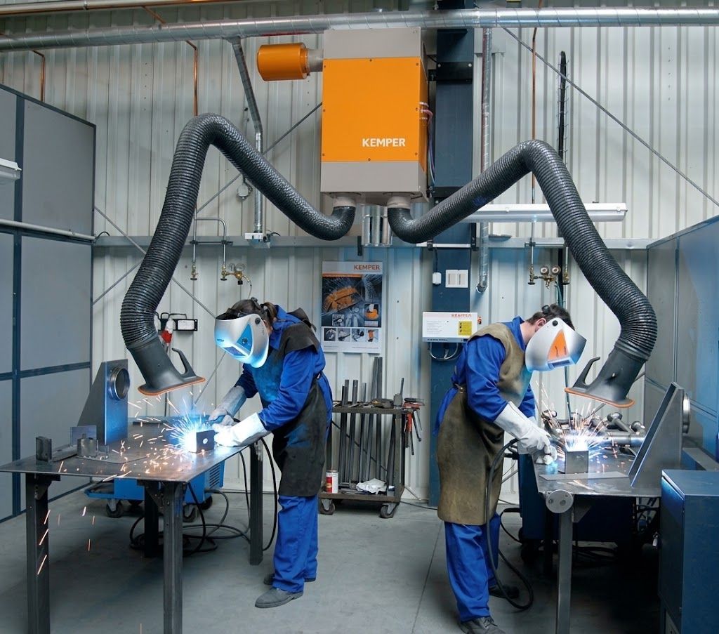Two workers in welding gear at metal workbenches under large extraction hoses in an industrial workshop