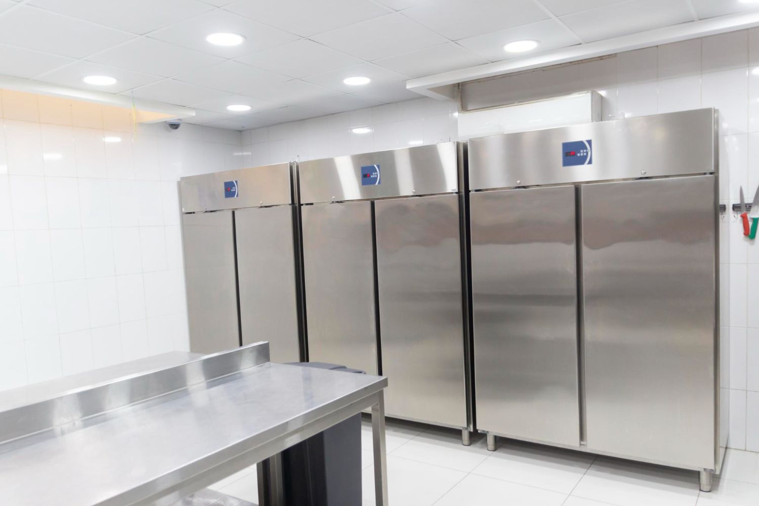 Three large, stainless-steel commercial refrigerators lined up along a white-tiled wall in a clean, professional kitchen.