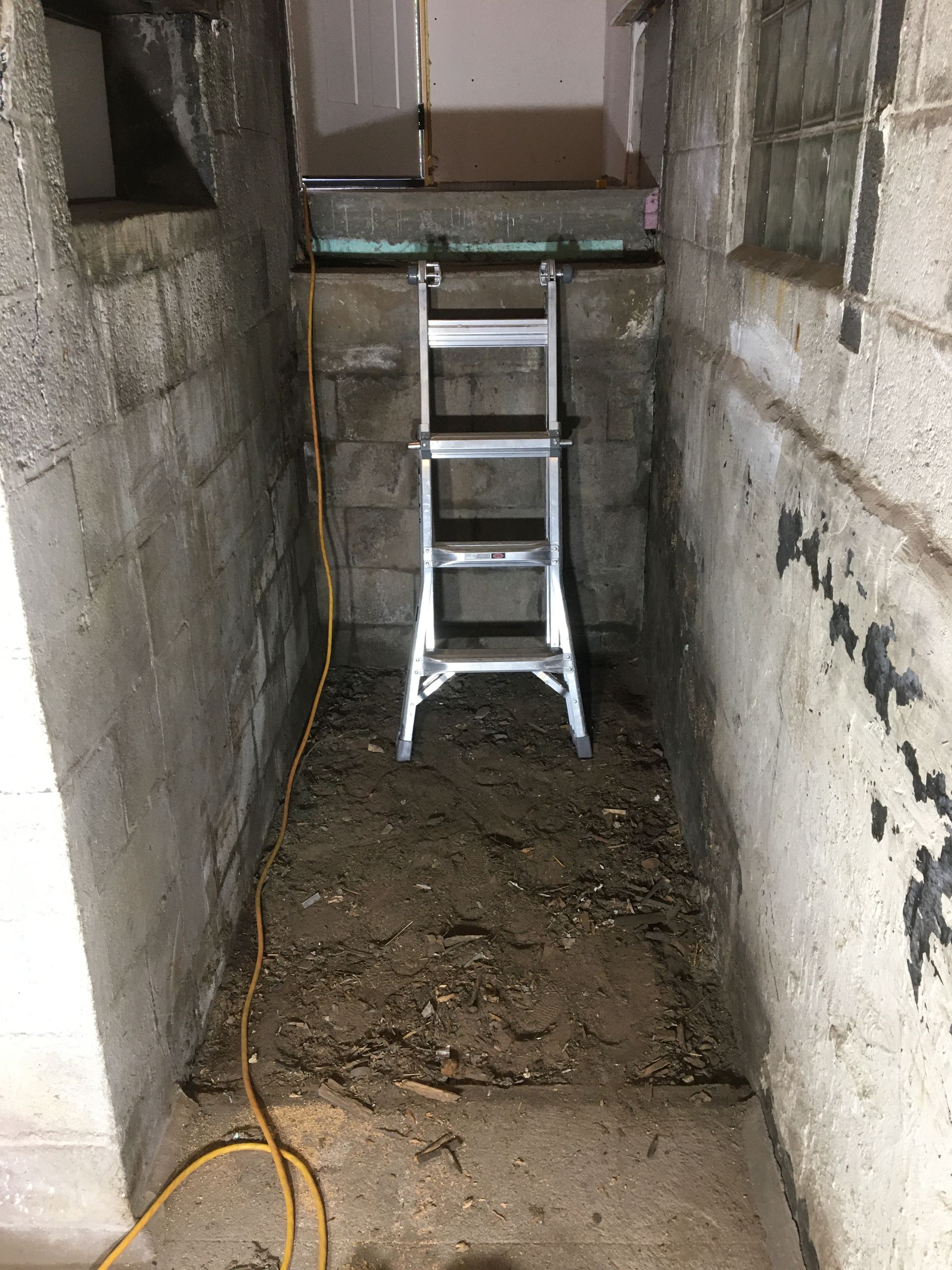Aluminum ladder in a narrow concrete trench with muddy floor and a cable on the ground.