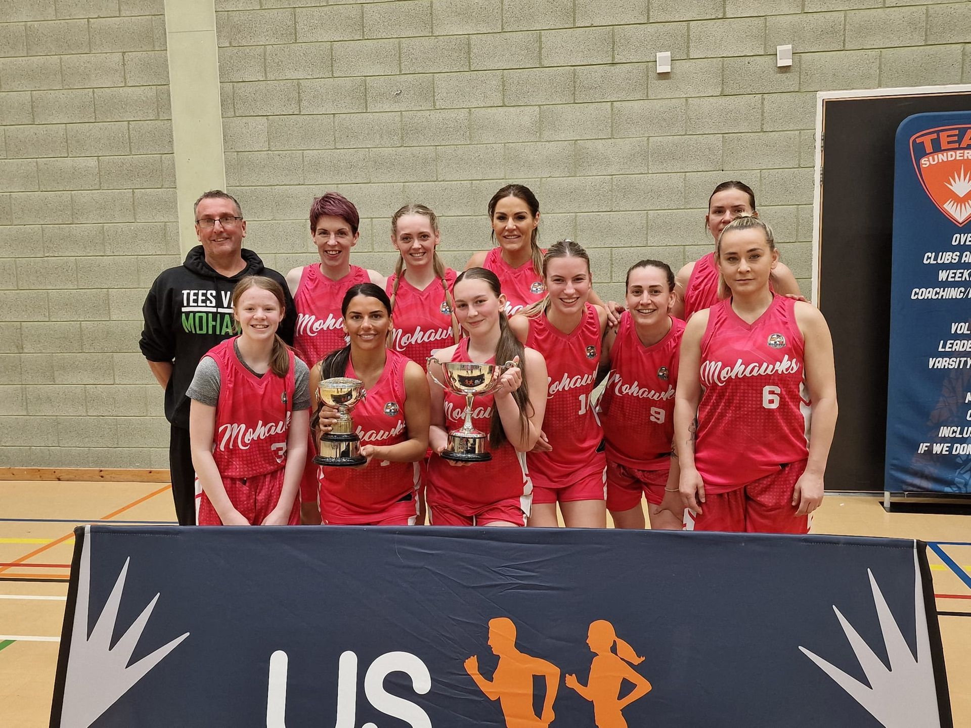 Volleyball team posing indoors behind a banner, holding a trophy and medals.