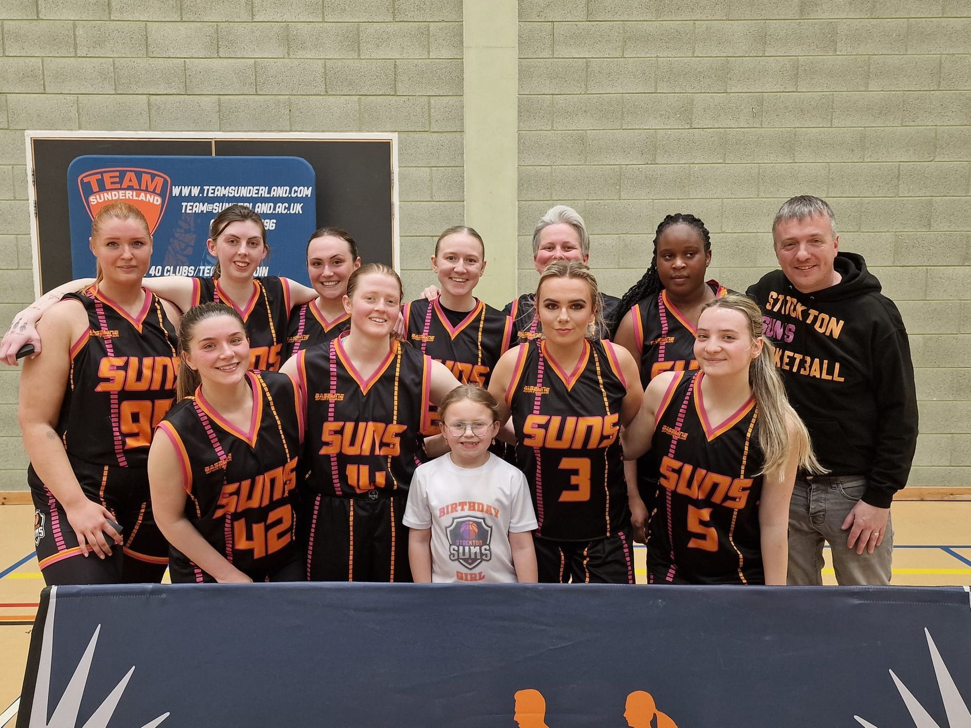 Basketball team posing outdoors in black and orange uniforms with a championship banner