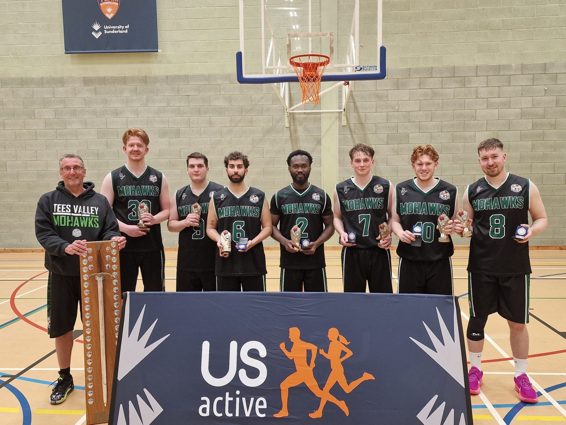 Eight men in black sports uniforms pose with trophies in a gym under a basketball hoop.
