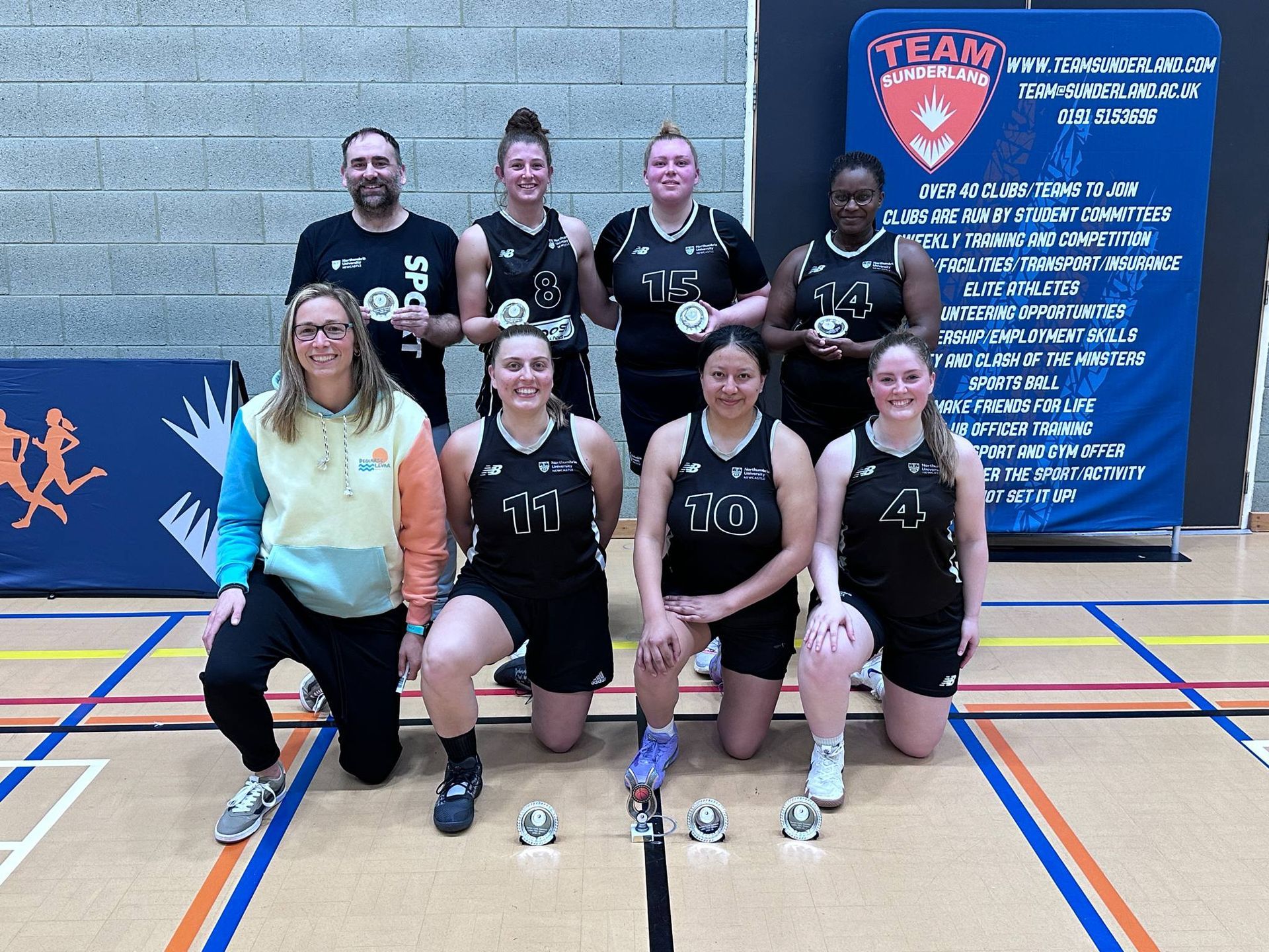 Indoor team photo of six athletes posing with medals on a gym floor, in front of a blue event banner