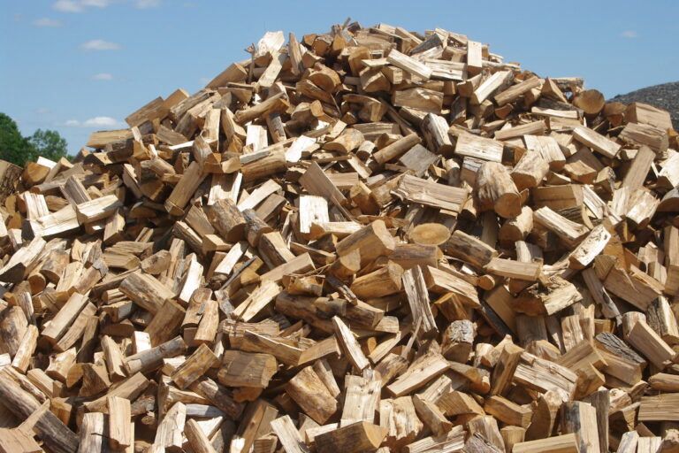 A large, cone-shaped mound of chopped, light brown firewood piled high against a blue sky.