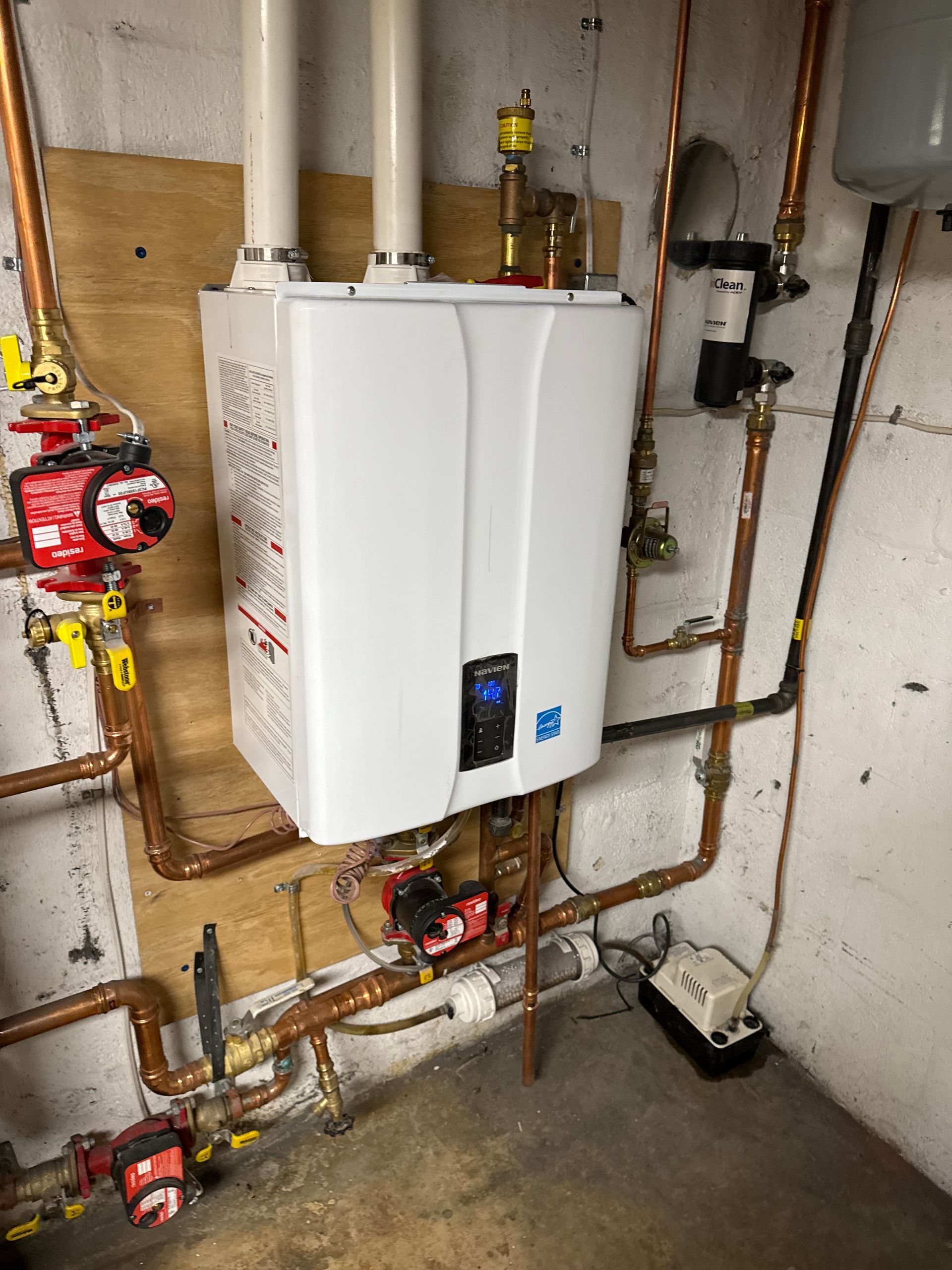 A wall-mounted white boiler with copper pipes in a basement setting. Two white exhaust pipes extend upwards. Red pumps and other plumbing are also visible.