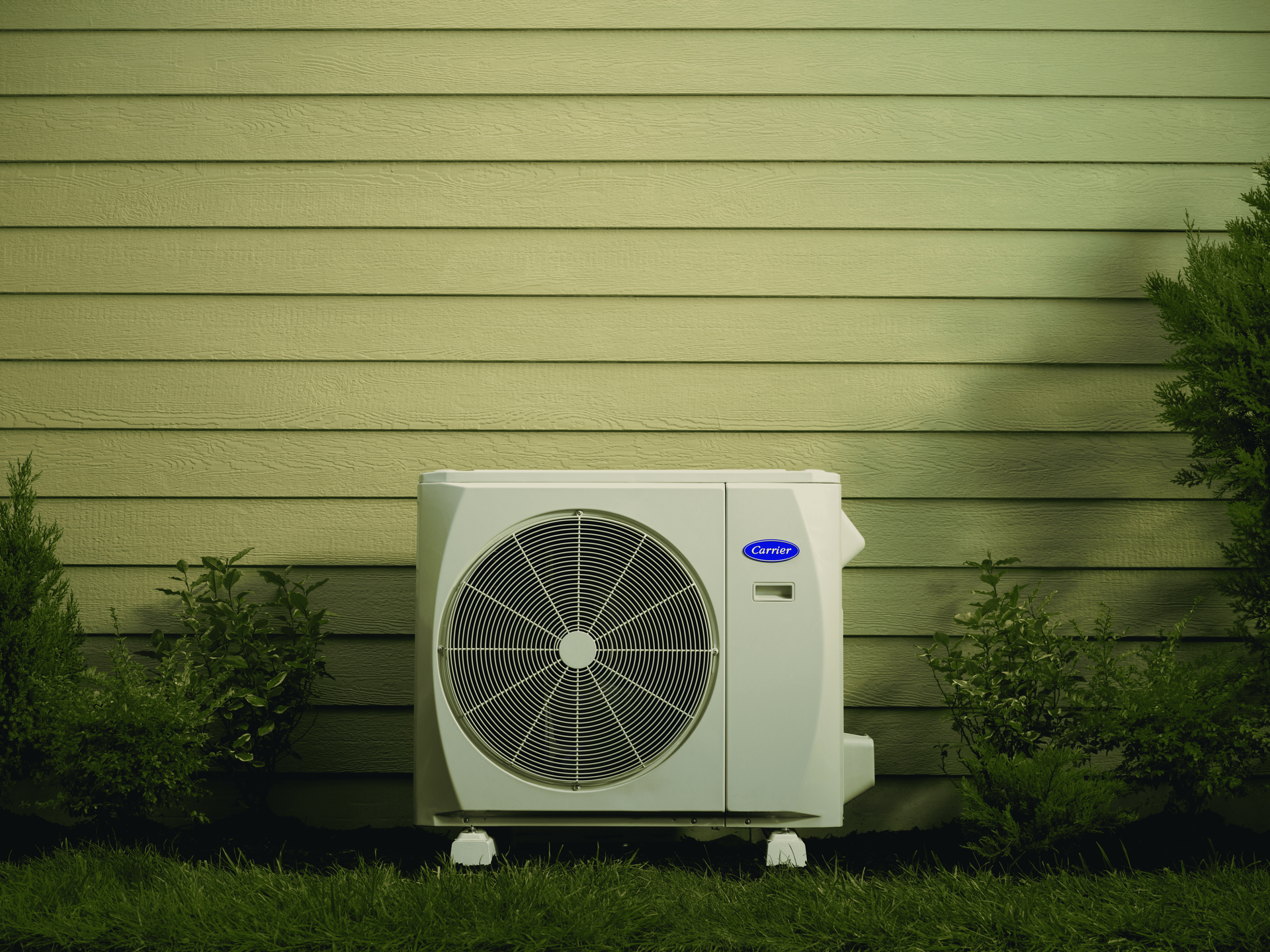 White outdoor air conditioning unit near a beige wall and green bushes.