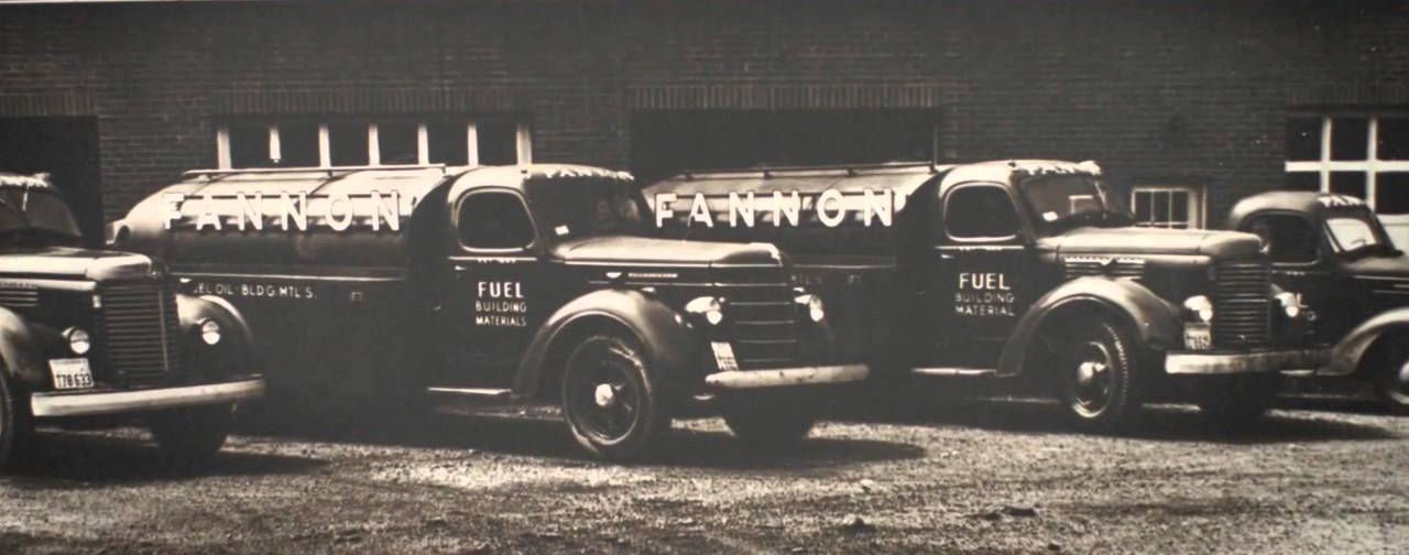 Black and white photograph of a row of tanker trucks parked in front of a building. The trucks have the name
