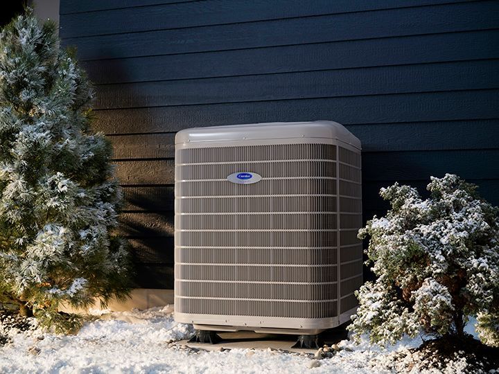 An air conditioning unit next to a house, surrounded by snowy bushes. Dark blue siding.