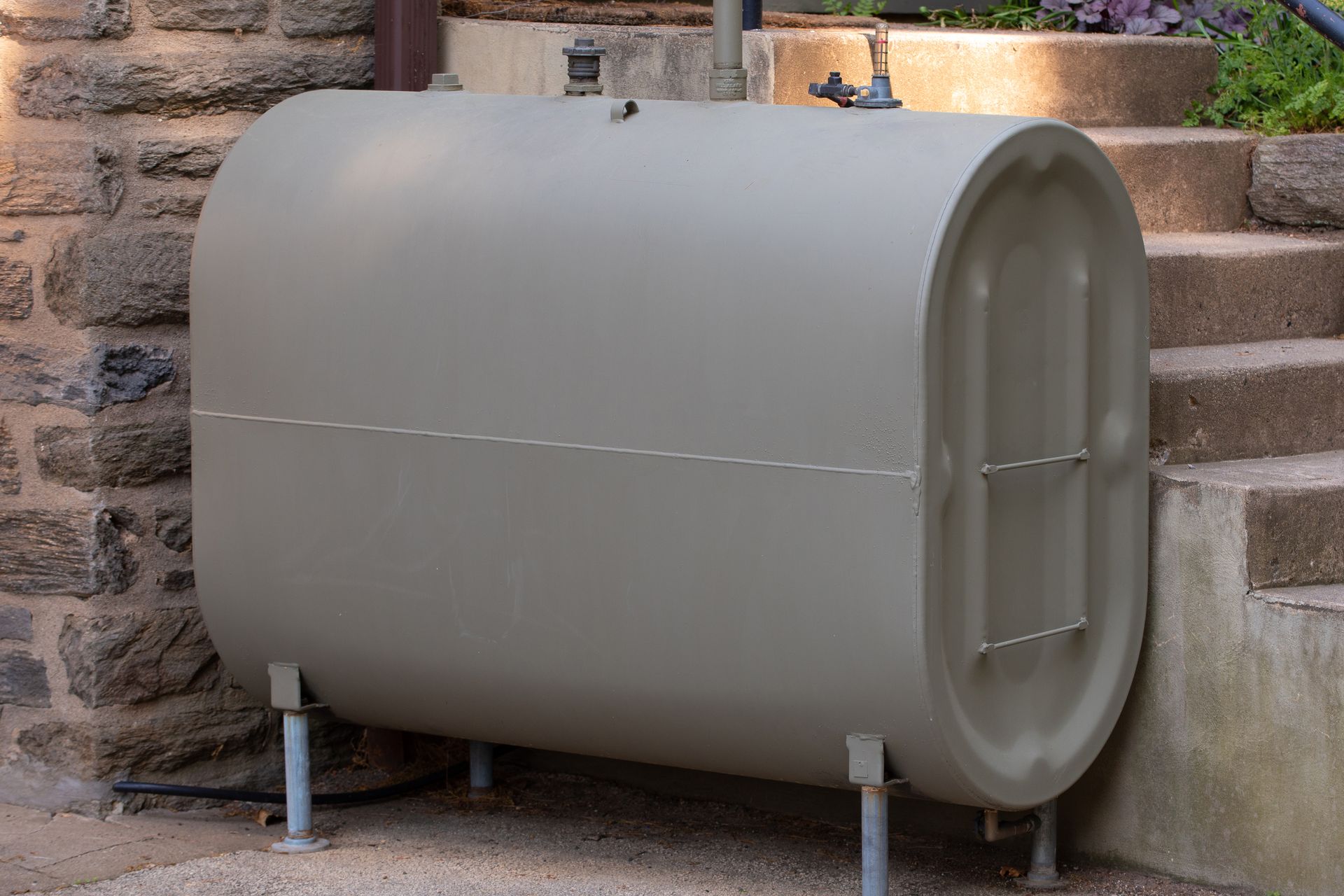 A horizontal, olive-green oil tank with metal legs, set against stone steps.