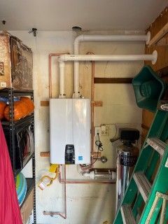 A white wall-mounted tankless water heater in a garage. Copper pipes connect to the heater, and a ladder leans nearby.