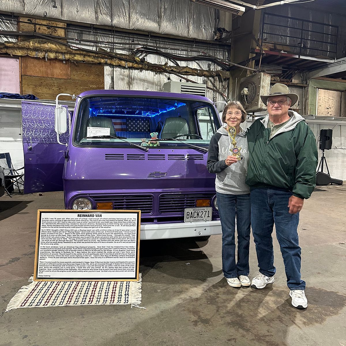 Two people standing beside an award-winning purple van in a warehouse, with a signboard in front at a classic car show