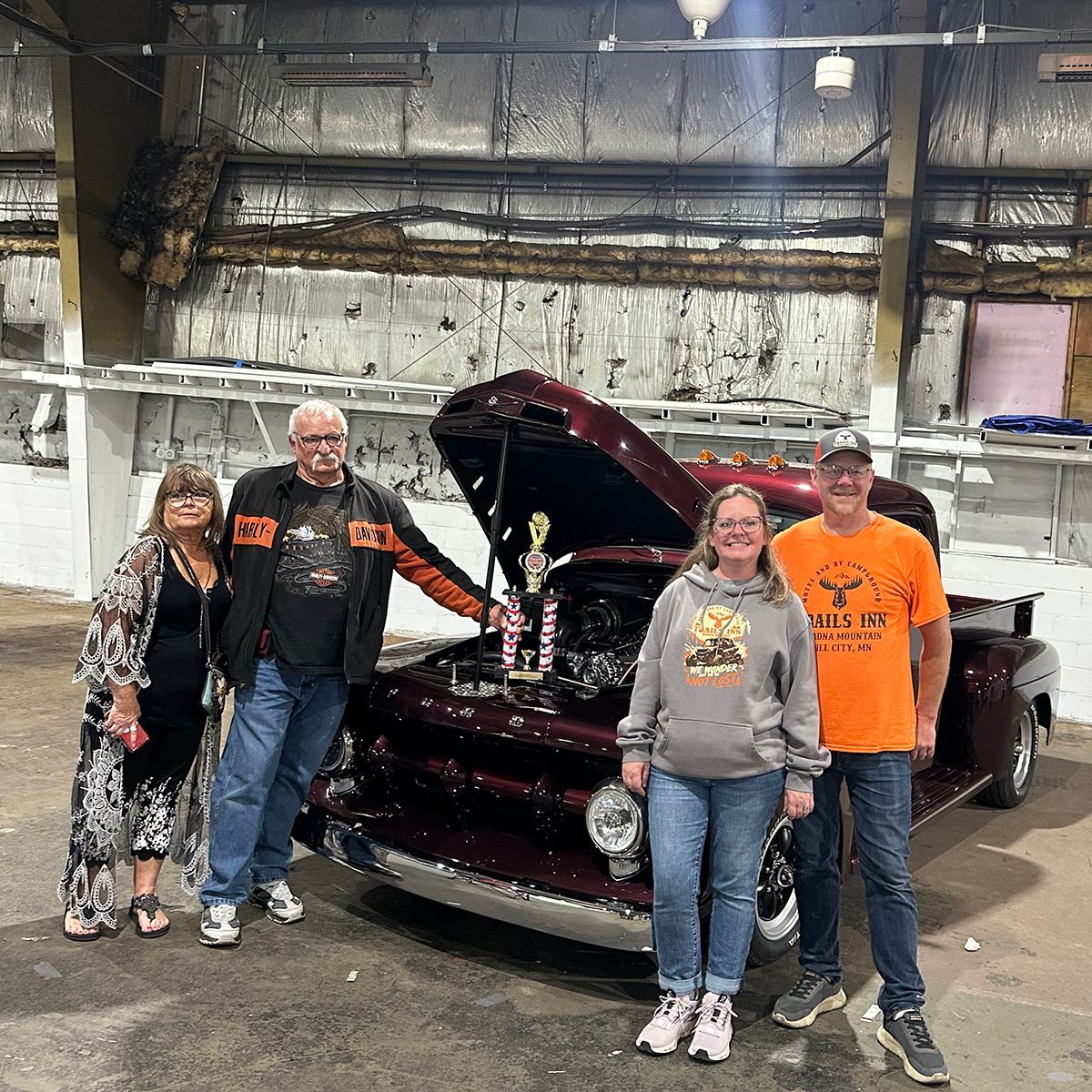 Four people pose beside an award-winning maroon classic car with an open hood inside a warehouse
