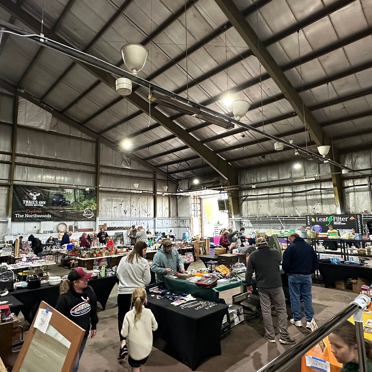 Indoor flea market with vendor tables, shoppers, and hanging lights in a large warehouse-like hall