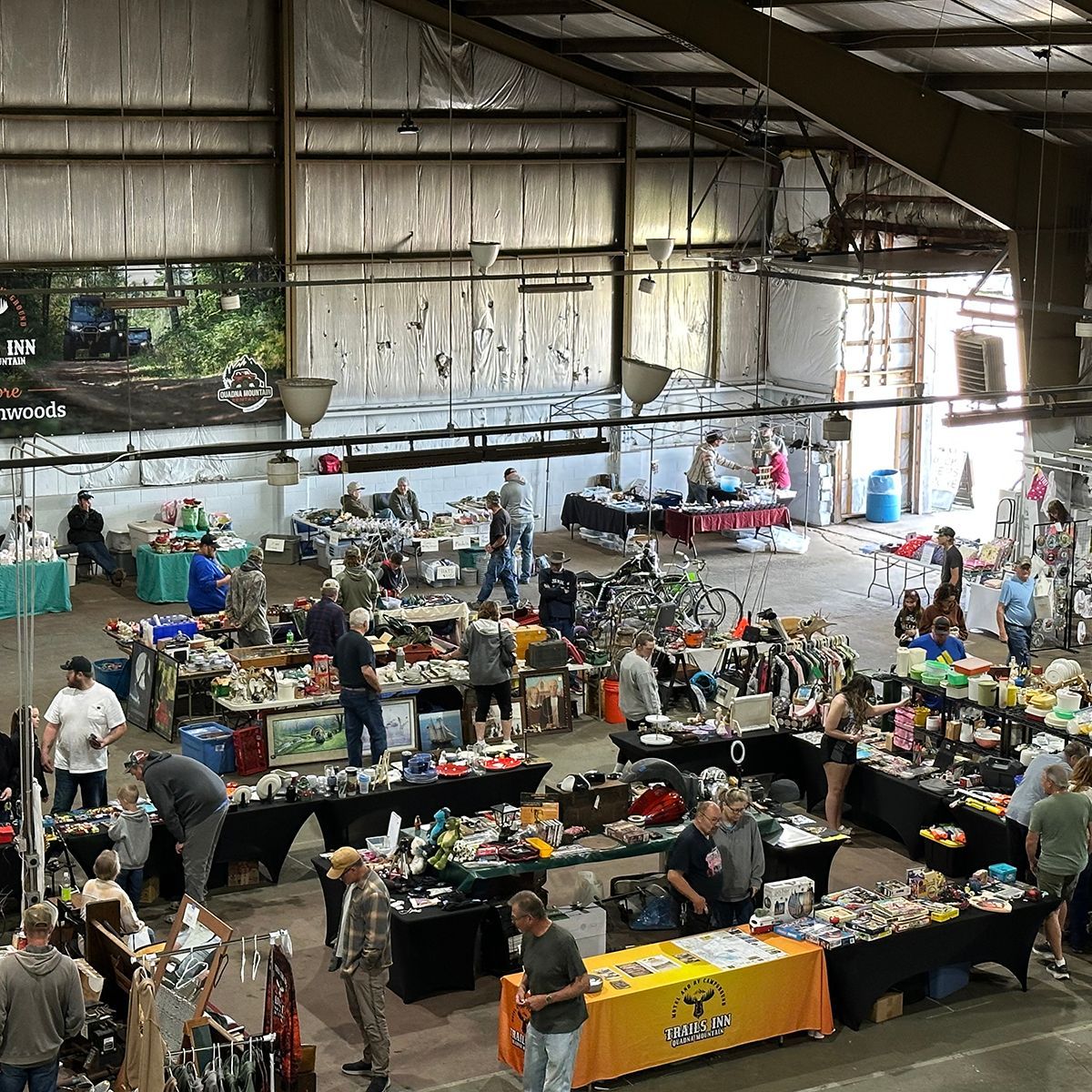 Indoor flea market with rows of tables, clothing racks, and shoppers under a large warehouse roof