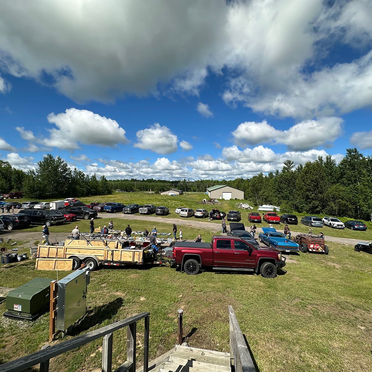 Rural outdoor event with parked pickup trucks, trailers, and scattered equipment under a cloudy blue sky