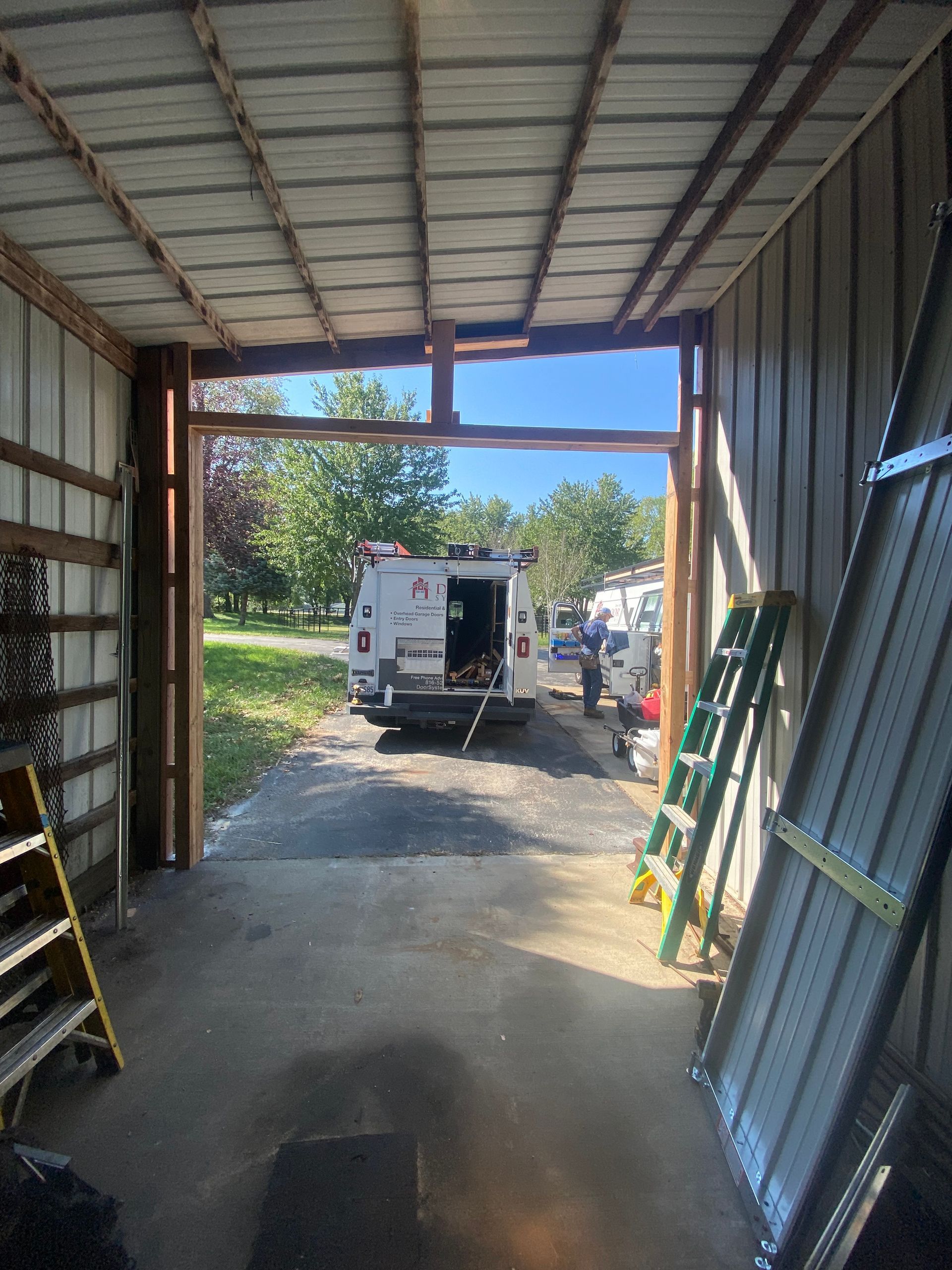 Inside a garage, a white utility van is parked outside an open doorway with ladders leaning nearby.
