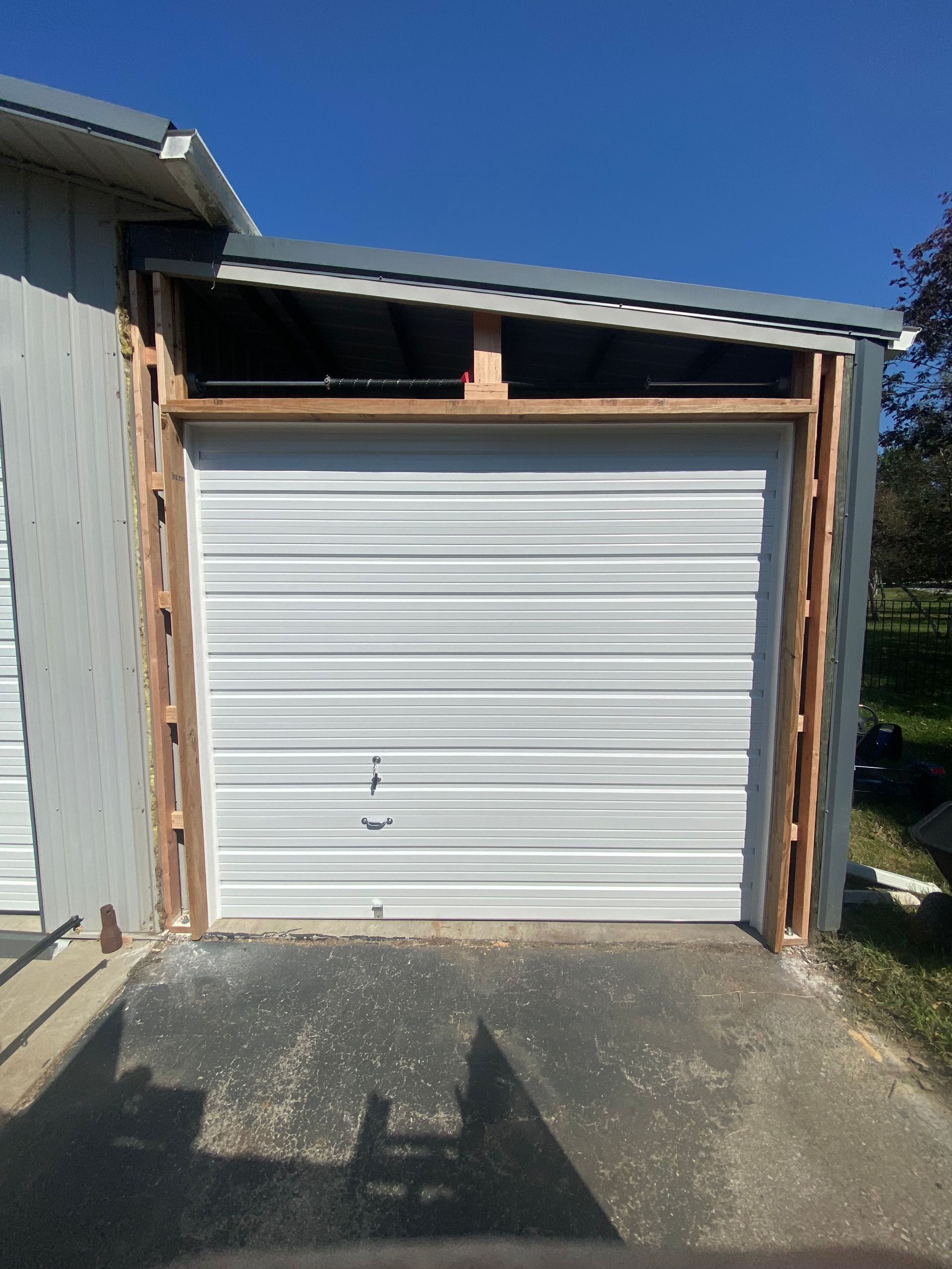 White roll-up garage door in a narrow wooden-framed carport beside a house under a clear blue sky