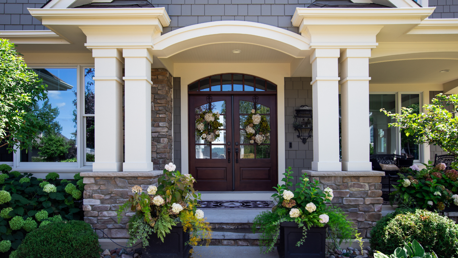 Front entrance of a house with white columns, stone base, dark wood double doors, and potted flowers