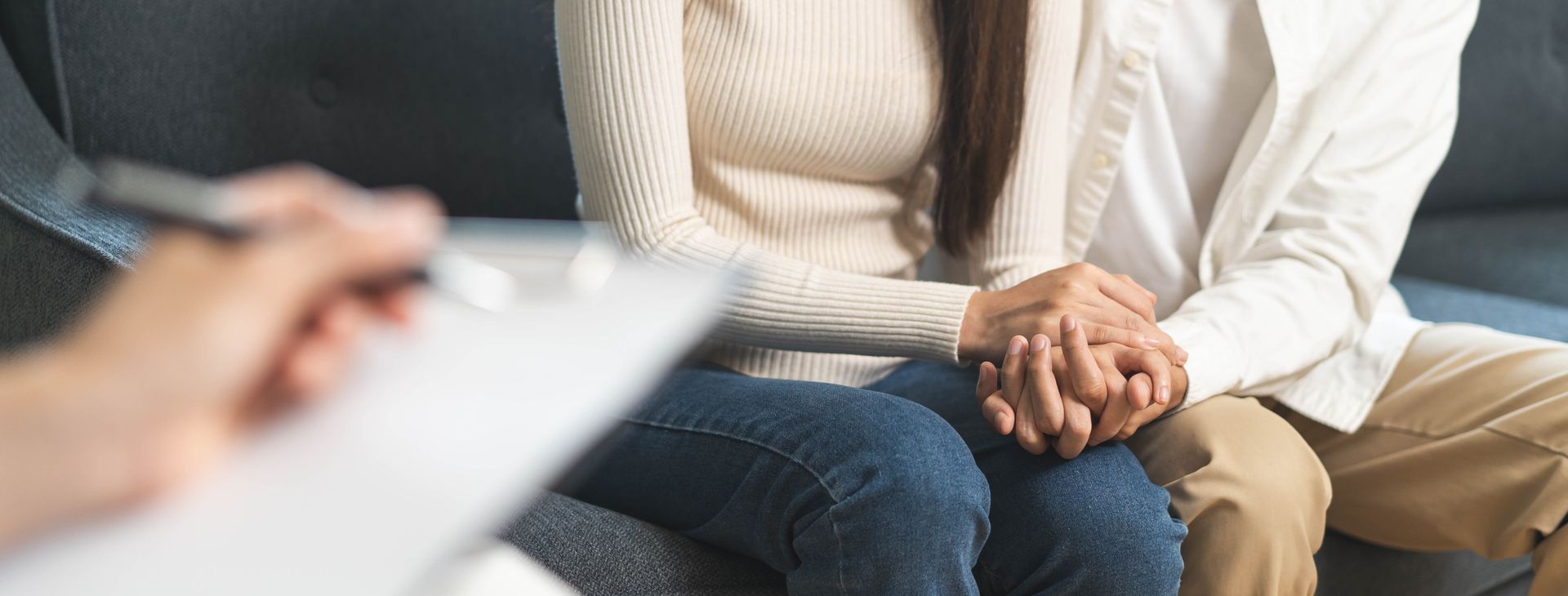 Couple holding hands during therapy session. Therapist's hand holding a pen and clipboard is in the foreground.