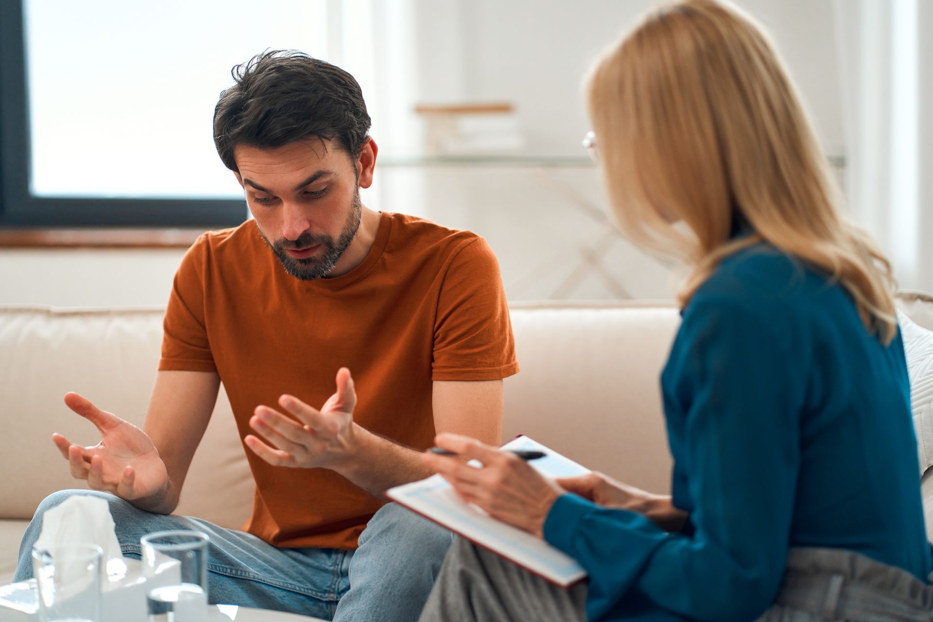 Man talking to a therapist; both seated on a couch. The therapist takes notes.