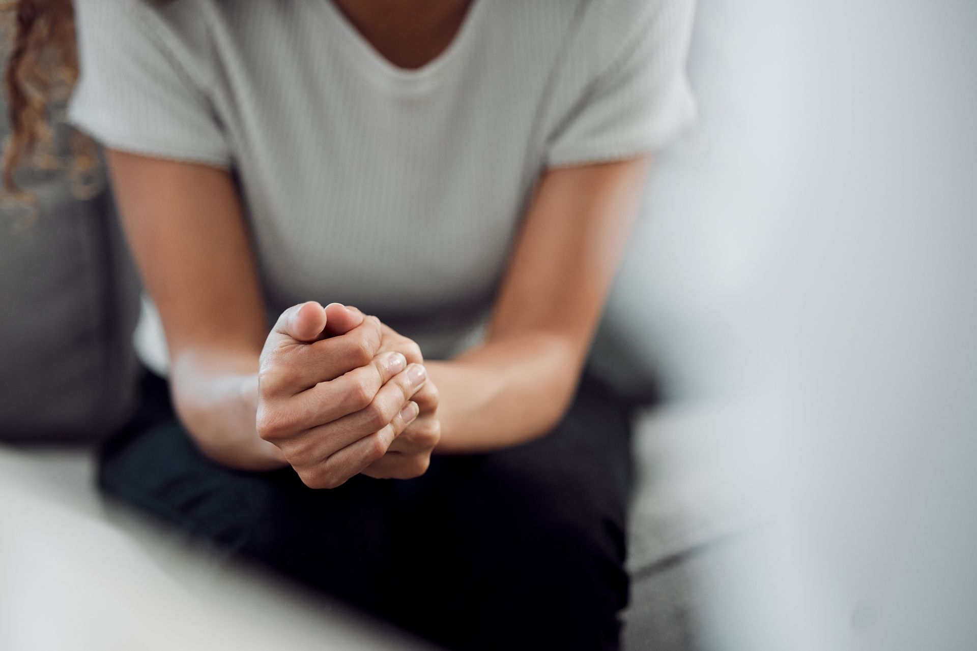 Person sitting with clasped hands, appearing thoughtful, in neutral-toned room. Person sitting with hands clasped, resting on their lap.