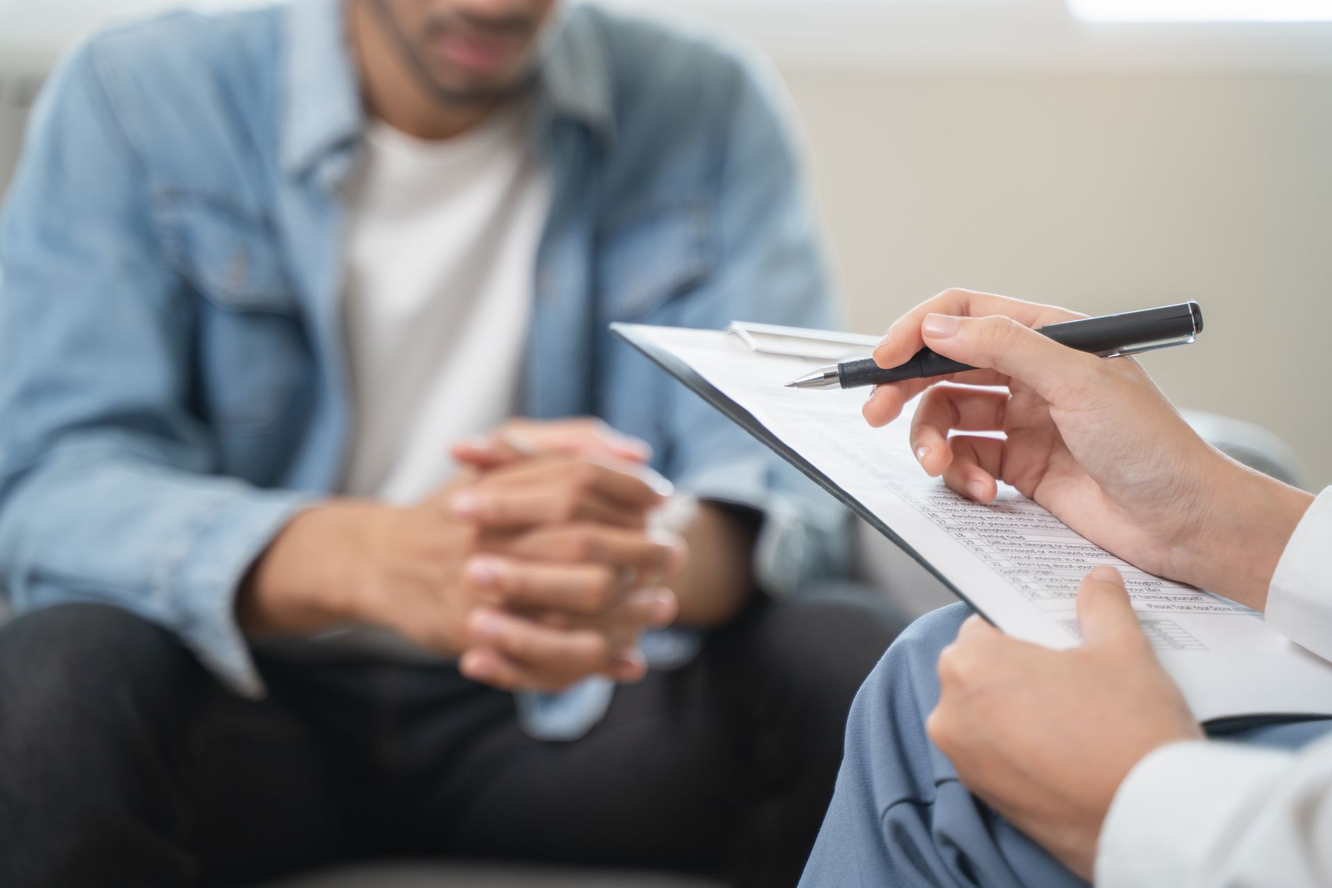 Person seated, hands clasped, being interviewed. A person holds a clipboard and pen. Person seated, hands clasped, being interviewed. A person holds a clipboard and pen.