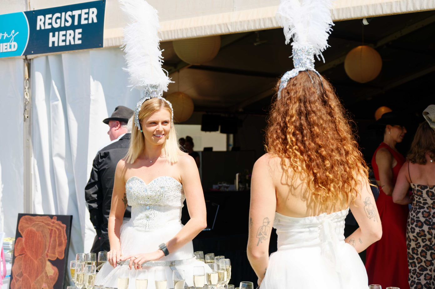 Two women in white dresses are standing in front of a sign that says register here