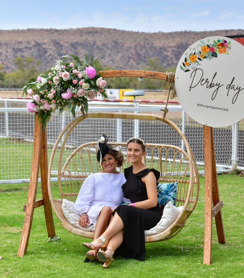 Two women are sitting on a wicker swing in a field.