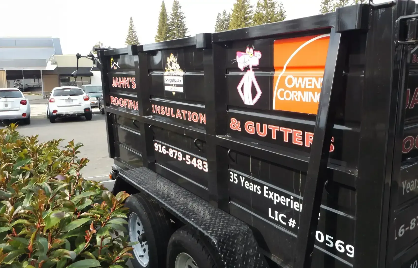 Black dump trailer with company logos parked in a lot beside cars and a building