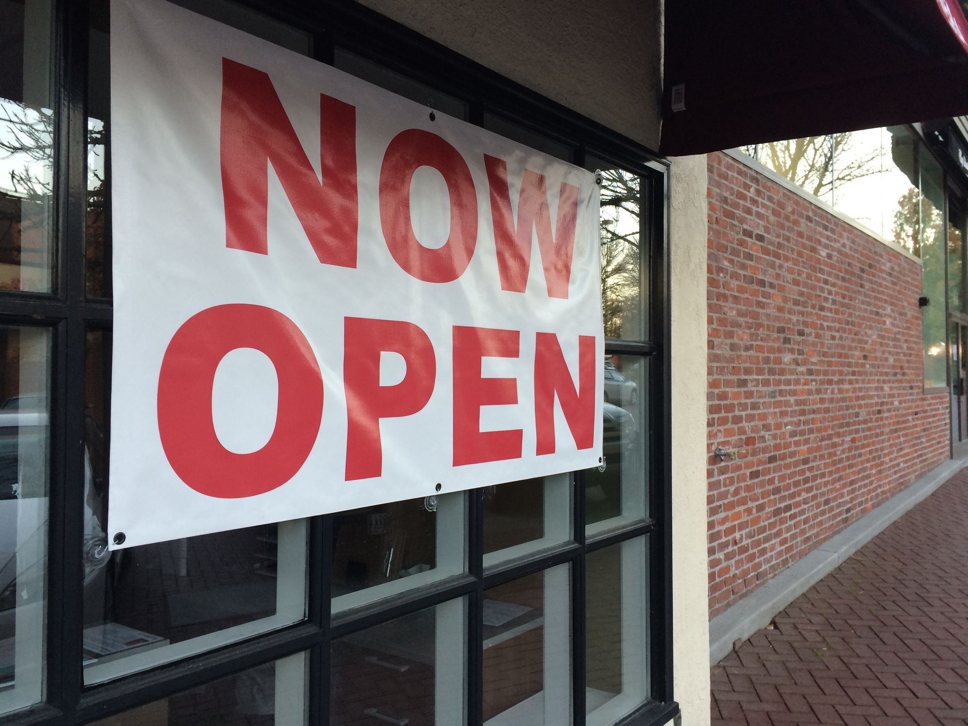“Now Open” sign in red letters hanging in a storefront window