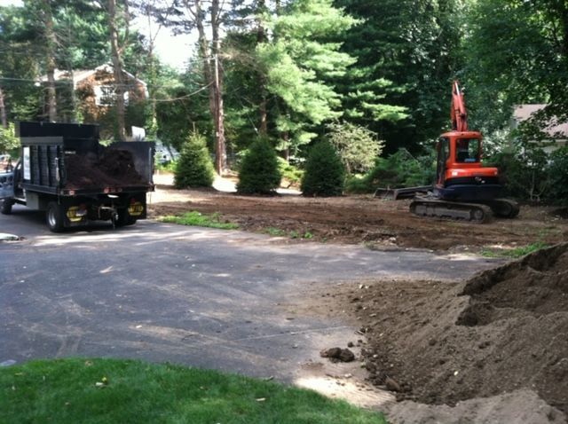 Construction equipment clearing a wooded driveway beside a gravel pile and truck