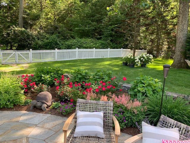 Patio chairs overlooking a lush garden with pink flowers, green lawn, and white fence in sunlight