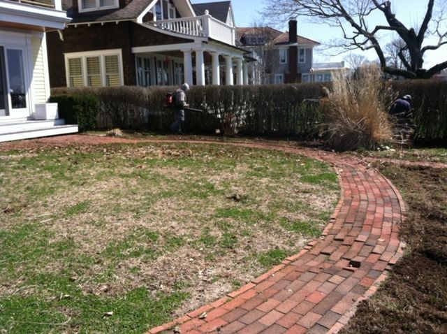 Curved brick walkway beside a house and hedge, leading through a grassy yard