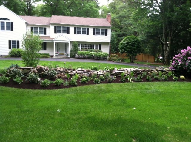 White two-story house with a front porch, landscaped flower bed, and green lawn in a wooded yard