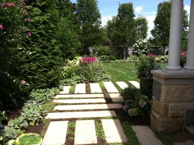 Stone garden path with white stepping stones leading through lush green shrubs and pink flowers under a sunny sky