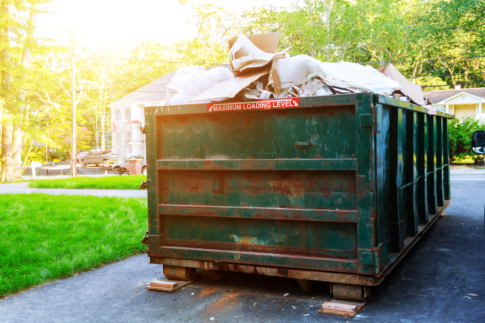 Large green dumpster filled with trash bags on a sunlit driveway beside a grassy lawn