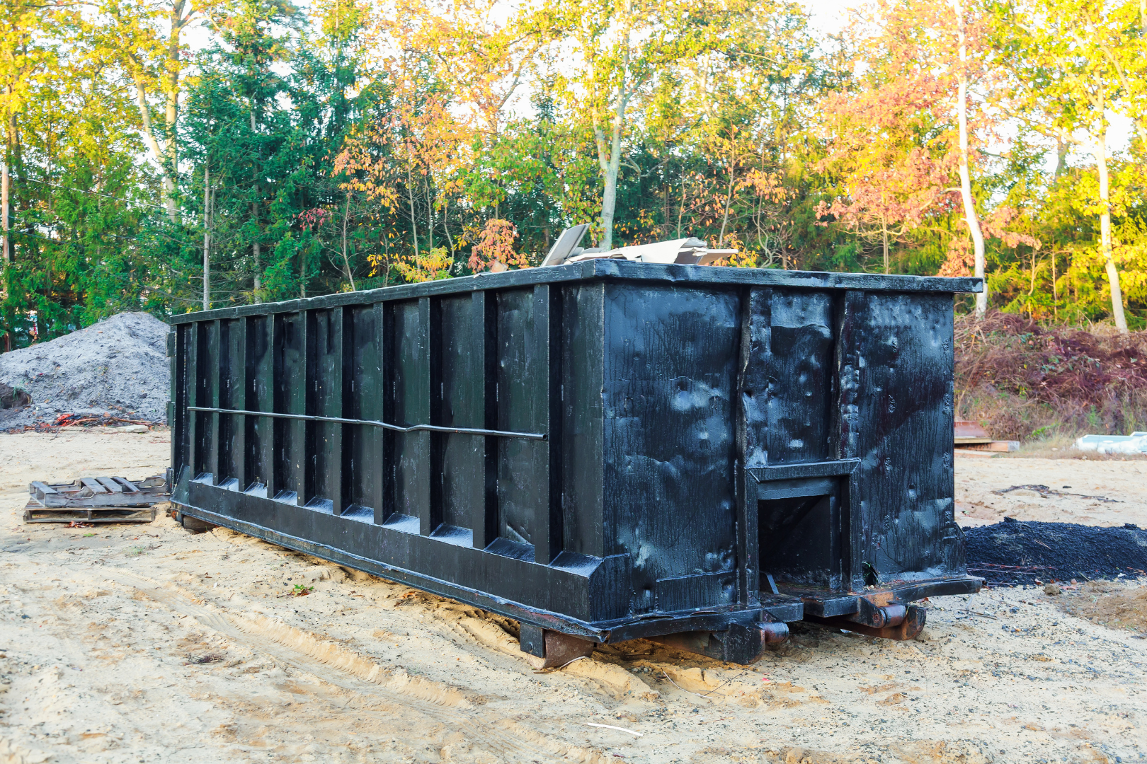 Large black dumpster container on dirt ground in a wooded outdoor area with autumn trees behind it