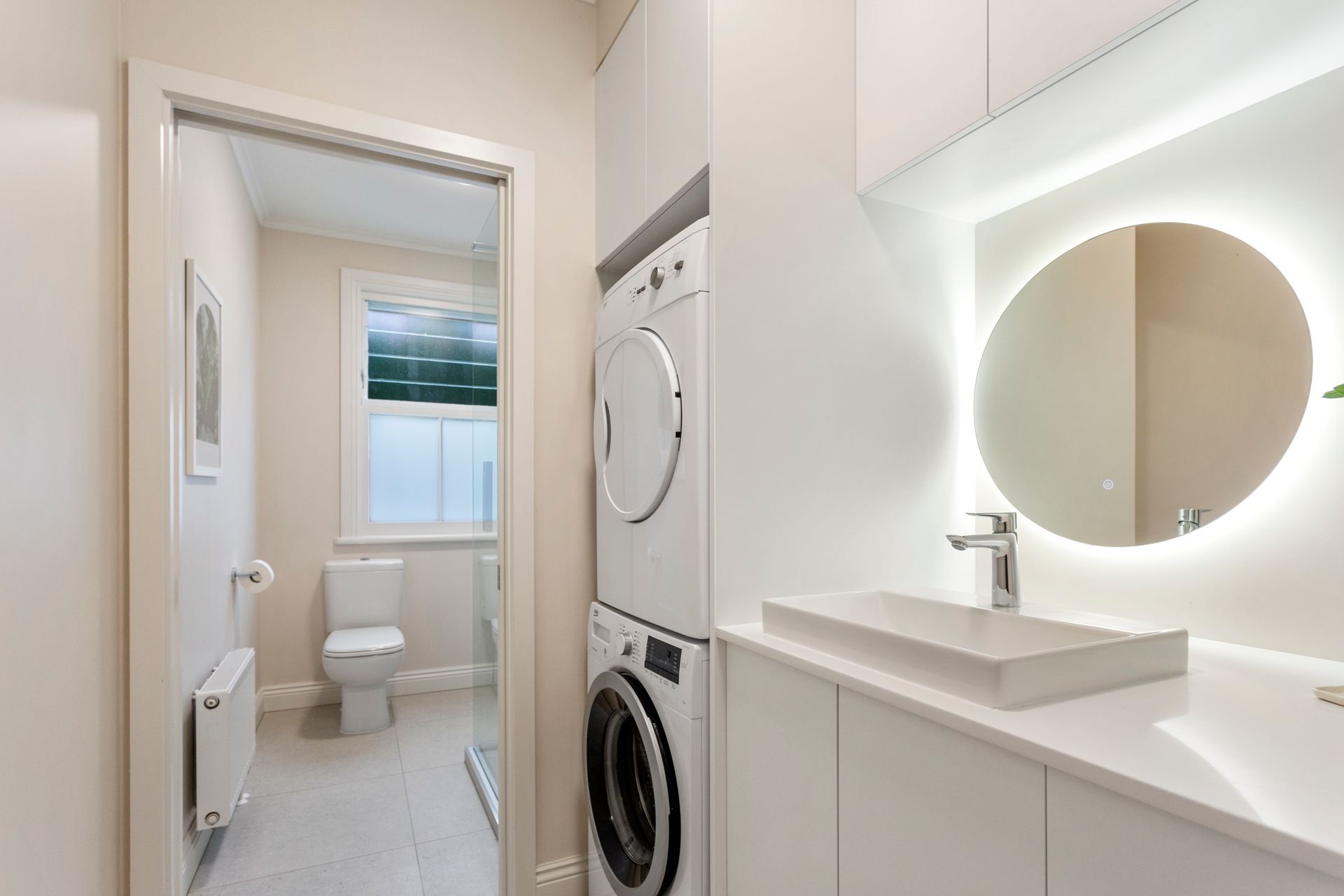 Small, modern laundry room with toilet, washer/dryer, sink, and backlit round mirror. White and neutral tones by Neda Builda