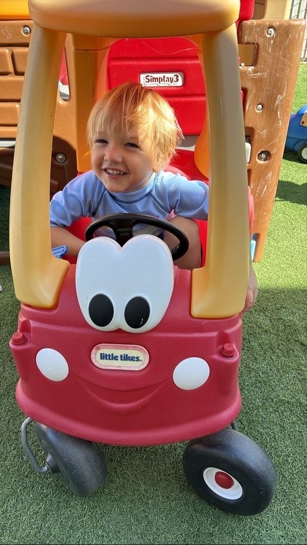 Child smiling in a red toy car with cartoon eyes outdoors on grass.