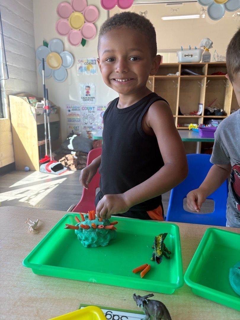 Smiling child in black shirt standing by green trays of toys in a classroom.