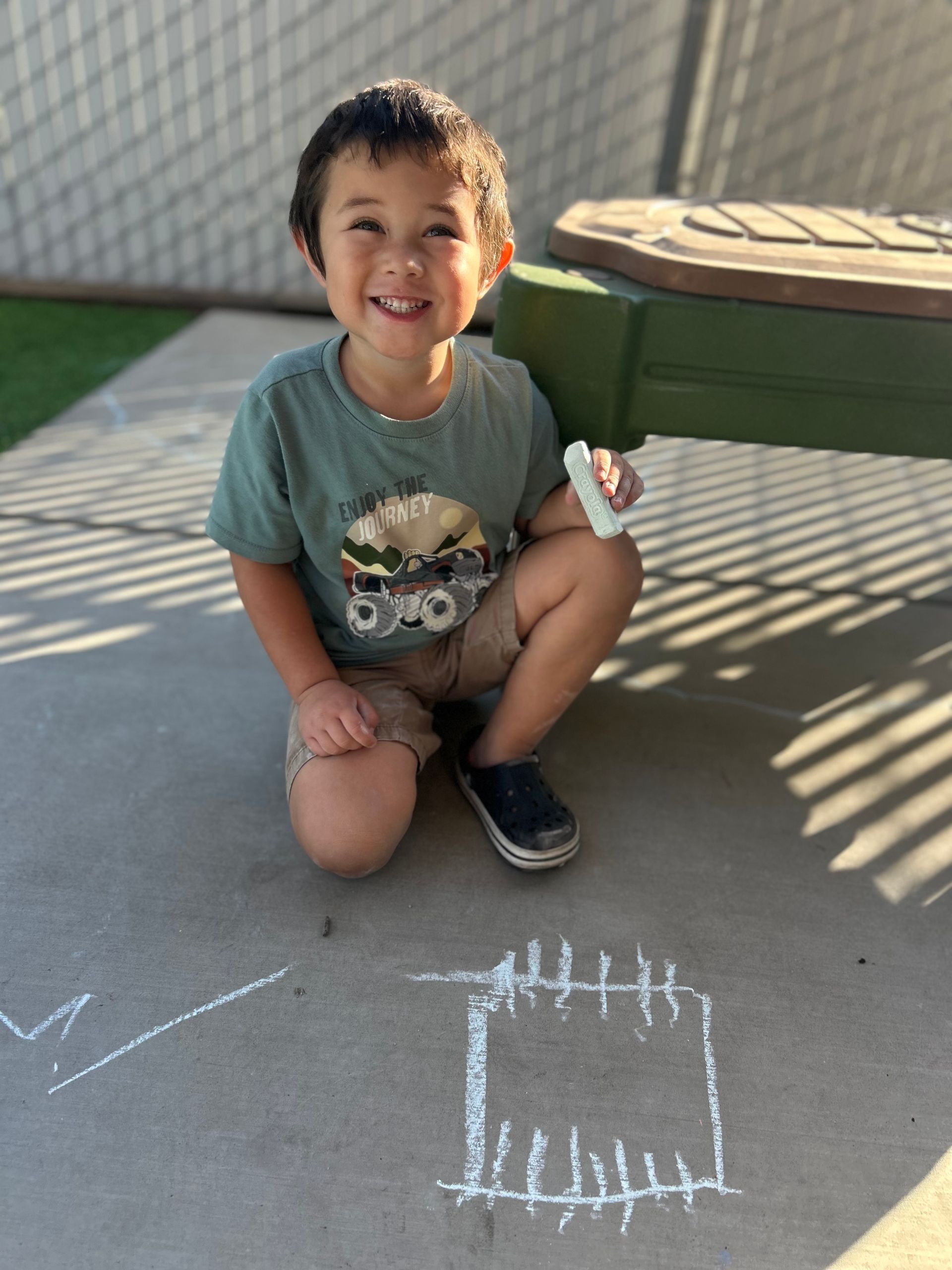 Smiling child crouches on a sunlit patio beside chalk drawings and a green storage bench.