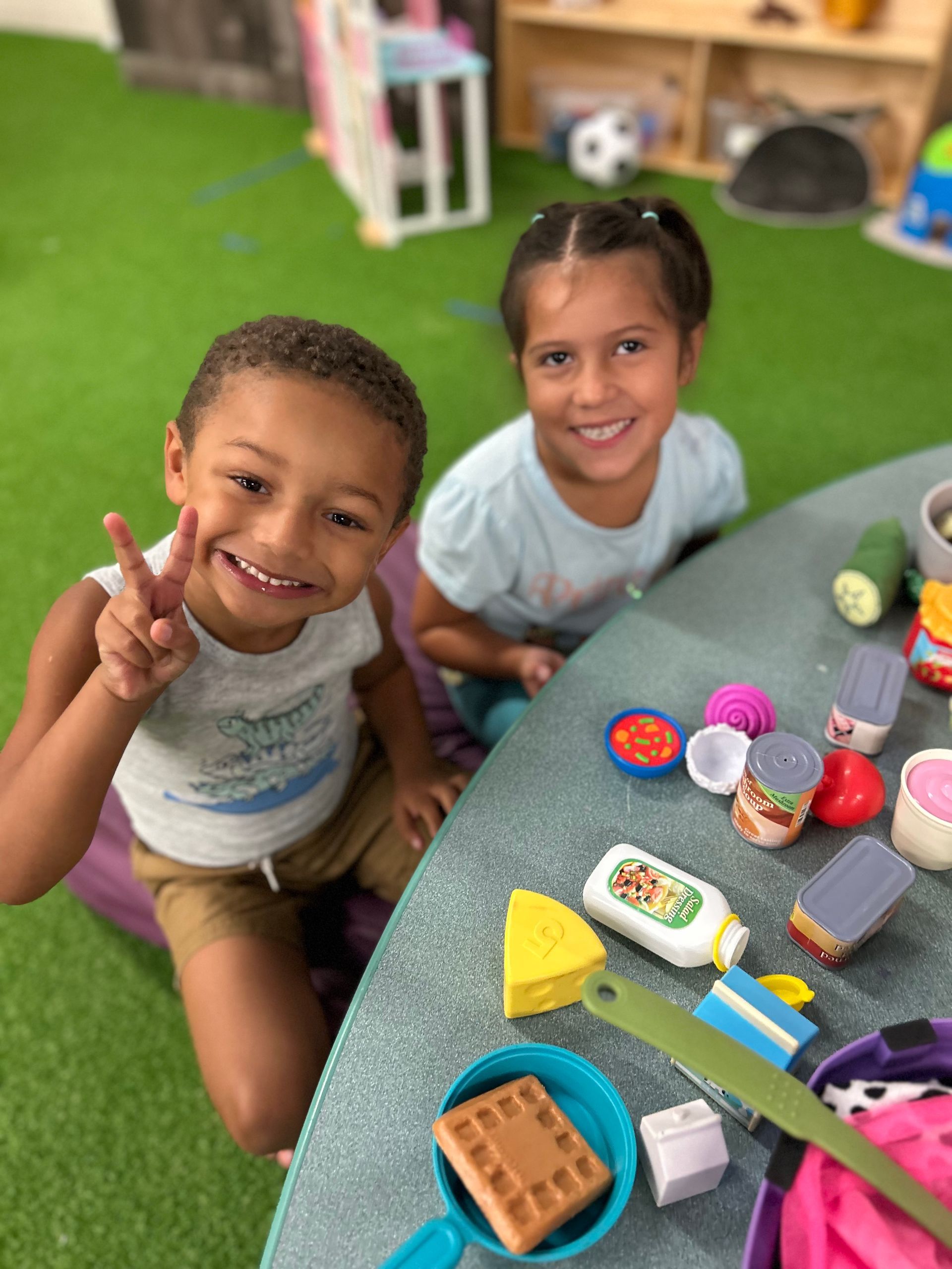 Two children smiling at a table with colorful toy food and kitchen playset pieces.