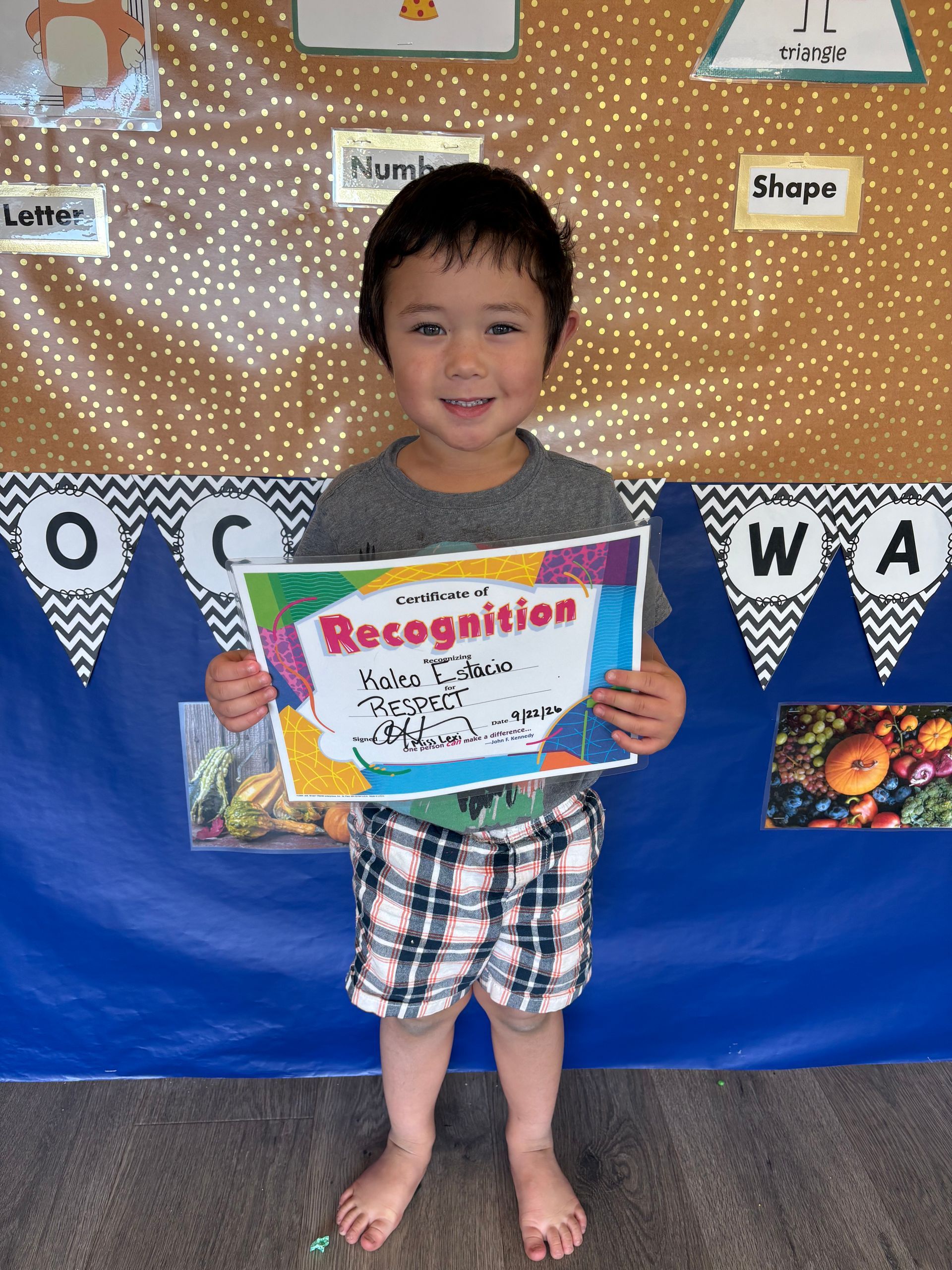 Child holding a colorful recognition certificate in a classroom display area, smiling and standing barefoot.