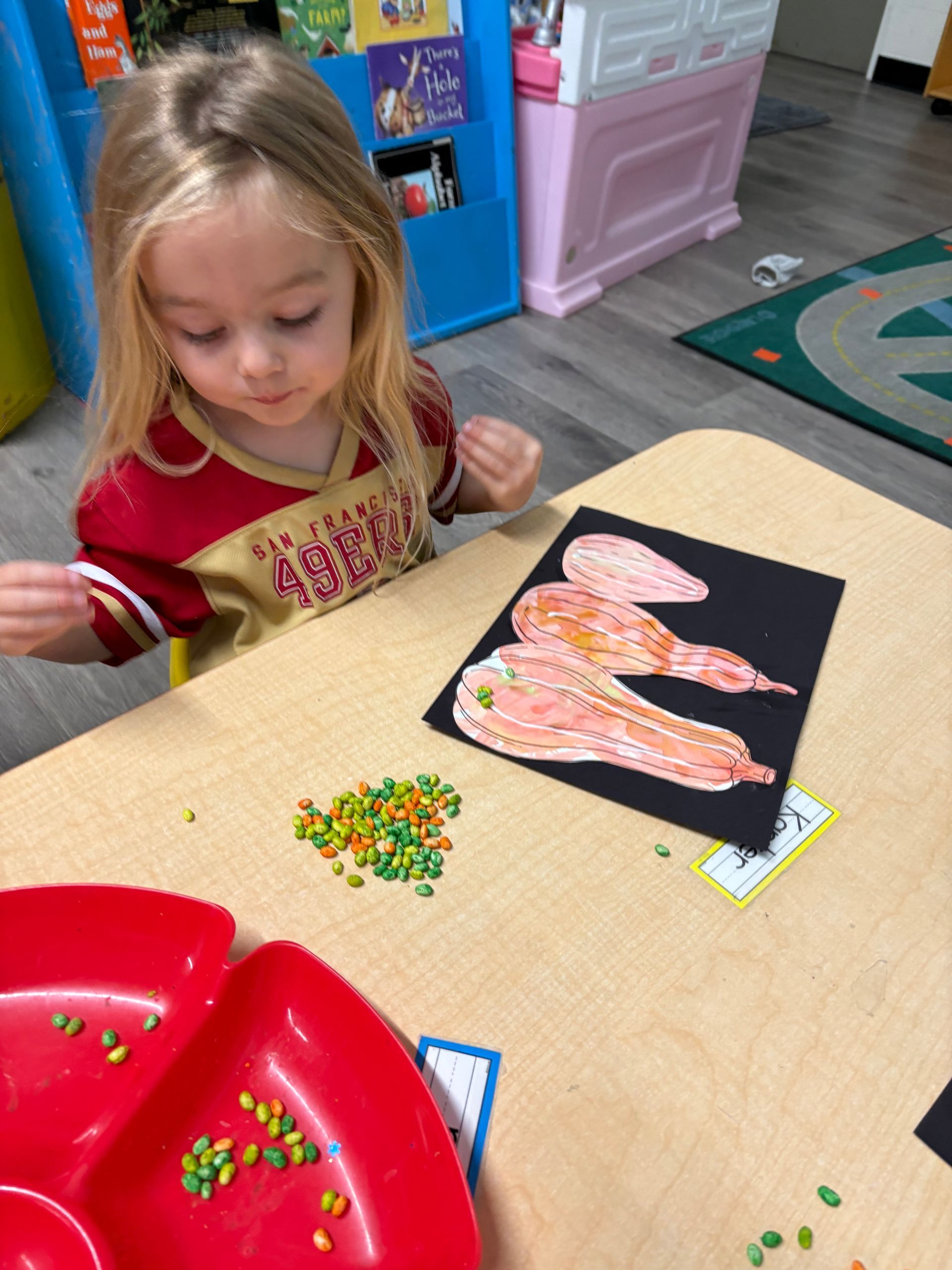 Child at a table sorting colorful beads onto a black paper shape in a classroom craft activity
