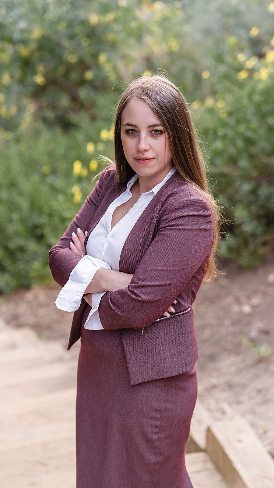 Woman in a burgundy suit stands outdoors with arms crossed, facing the camera.