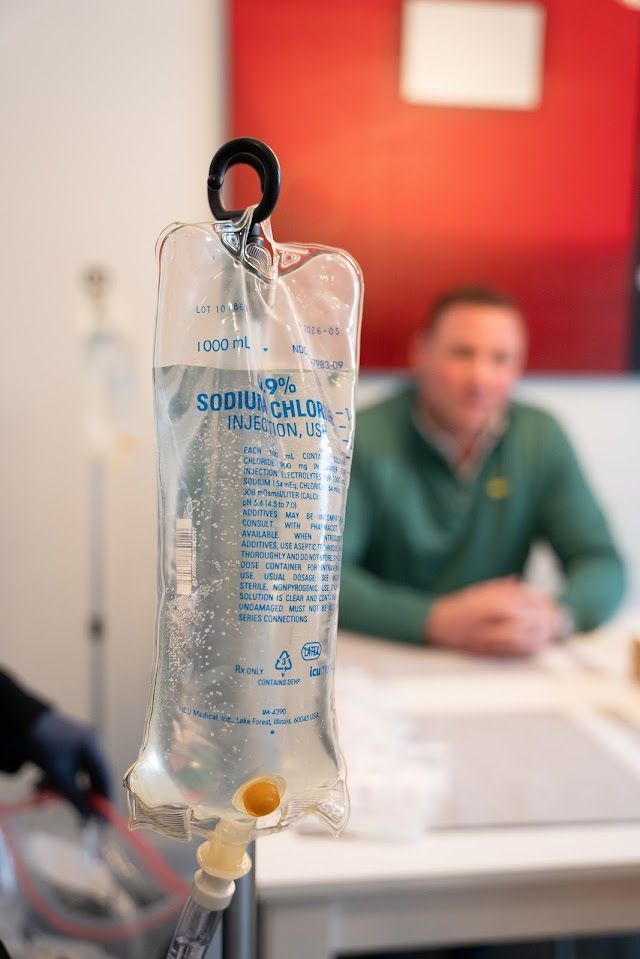 A man is sitting at a table with a bag of IV liquid in front of him.
