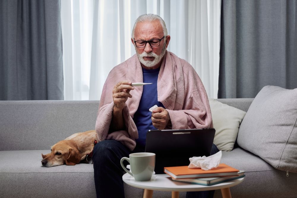 An elderly man is sitting on a couch holding a thermometer and a laptop.