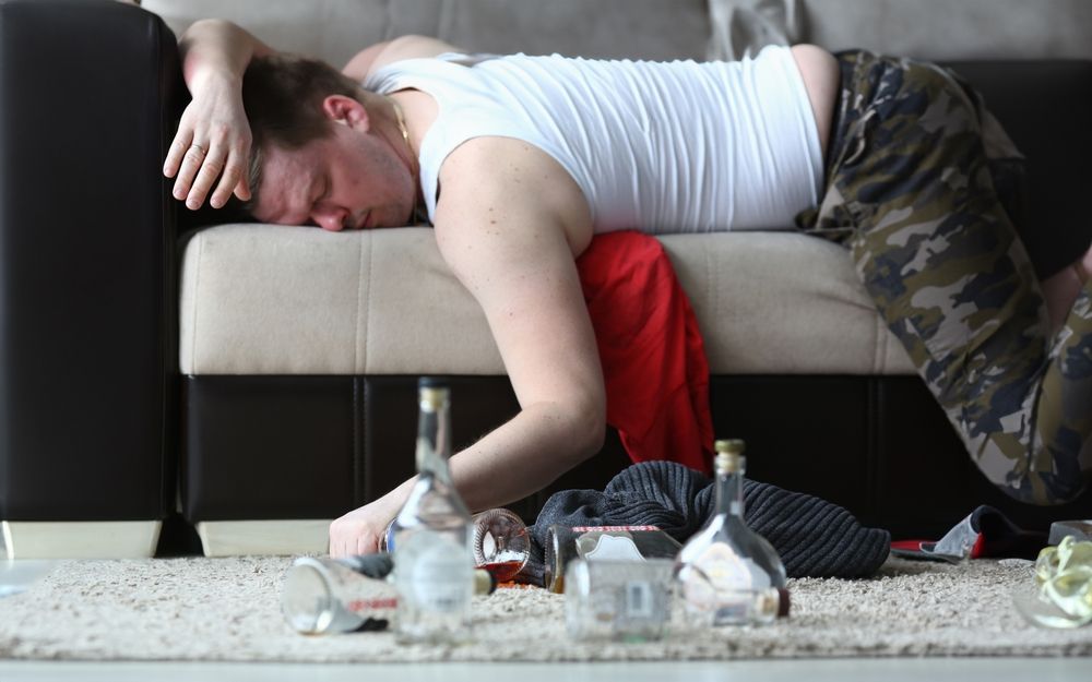 A man is laying on a couch with bottles of alcohol on the floor.